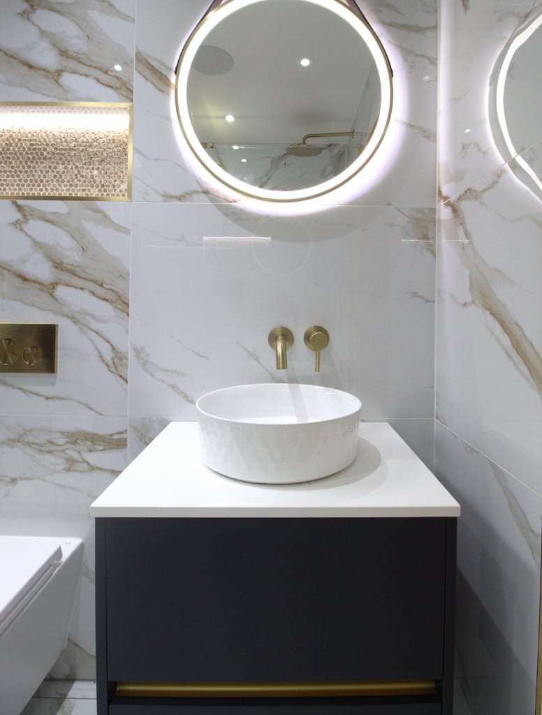 Sophisticated bathroom vanity in Hillington, Glasgow, with a round backlit mirror, white countertop basin, brass fixtures, and marble-style wall tiles for an opulent touch.