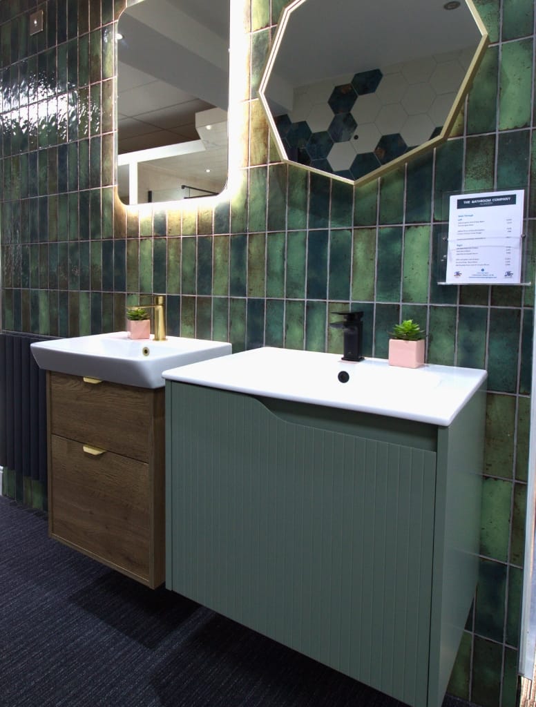Two vanity displays in a Glasgow bathroom showroom, featuring green and wood textures, hexagonal mirrors, and glossy green wall tiles for a stylish and earthy look.